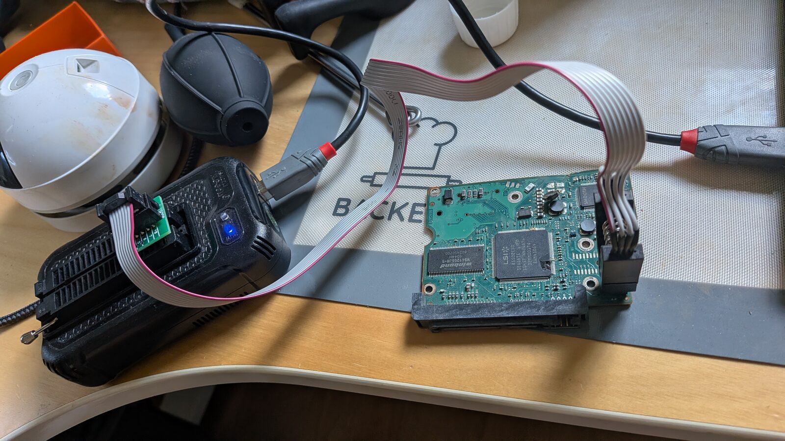 Workbench setup: a USB programmer with a SOIC8 clip attached via ribbon cable to a hard drive PCB, next to a rubber air blower bulb and other tools